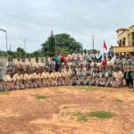 Group of schoolgirls in beige and green uniforms posing for a class photo outdoors with teachers and police officers in the background at a school yard